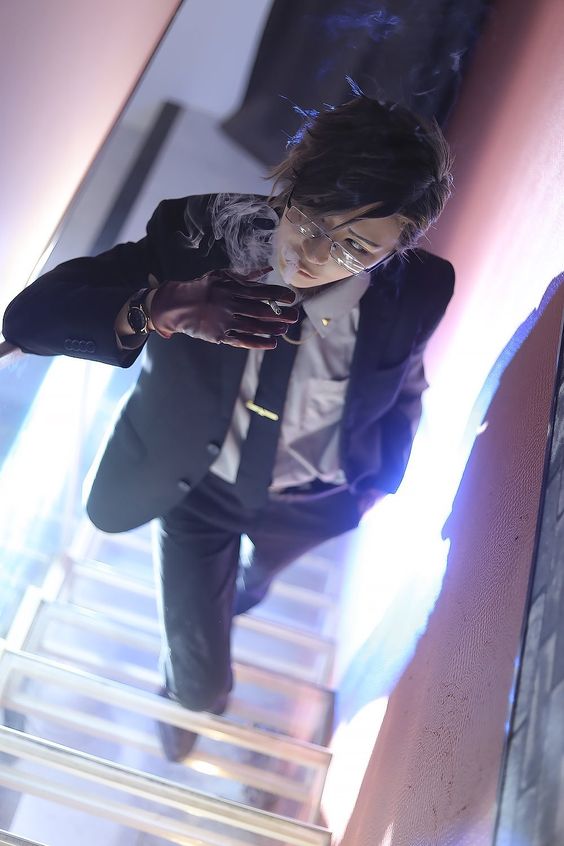 a man dressed in a suit and glasses is descending a staircase. he's smoking a cigarette and looking down towards the camera. the lighting is dramatic, with blue hues highlighting the scene from below. the perspective is from a low angle, creating a worm's eye view.