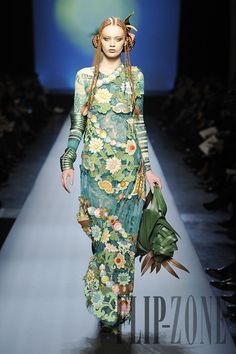 a model walks the runway in a green and blue floral dress. she is wearing a floral headband and a green bag. the model is walking on a light blue runway with other models in the background. the event is a fashion show.