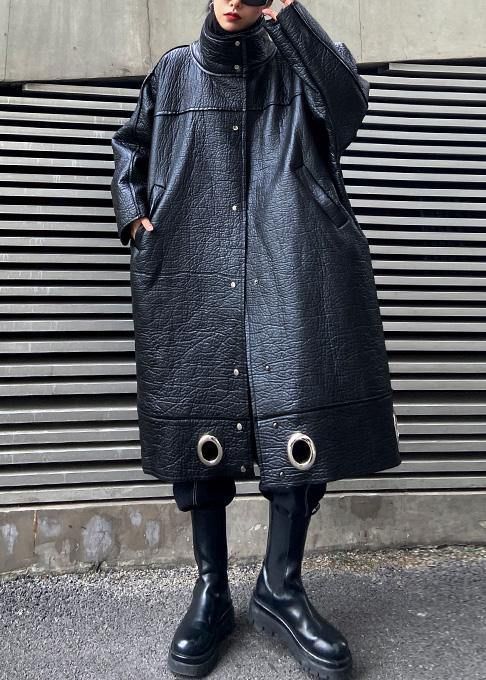 a woman wearing a long black leather coat and matching black boots stands in front of a black metal wall.  the coat has a relaxed fit and is decorated with silver grommets. the woman is looking at the camera and appears to be stylish and confident.