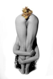 the image is a black and white artistic photograph of a nude woman. she is posed in a crouched position with her legs and arms wrapped around her body.  a small cluster of what appears to be dried flowers or blossoms rests on her buttocks. the overall feeling is one of sensuality and artistic expression. the monochrome palette emphasizes the form and texture of the body.