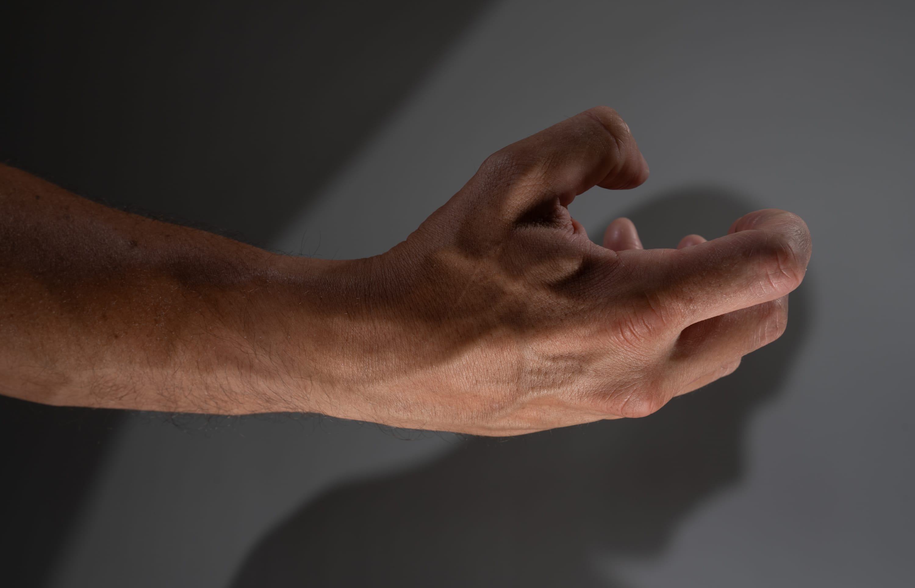 the image shows a man's hand and forearm extended towards the right side of the image.  the hand is partially curled and the fingers are slightly spread apart.  the arm is muscular and the skin is tanned.  there is a shadow of the hand and arm cast on the wall behind it.  the background is a neutral gray.