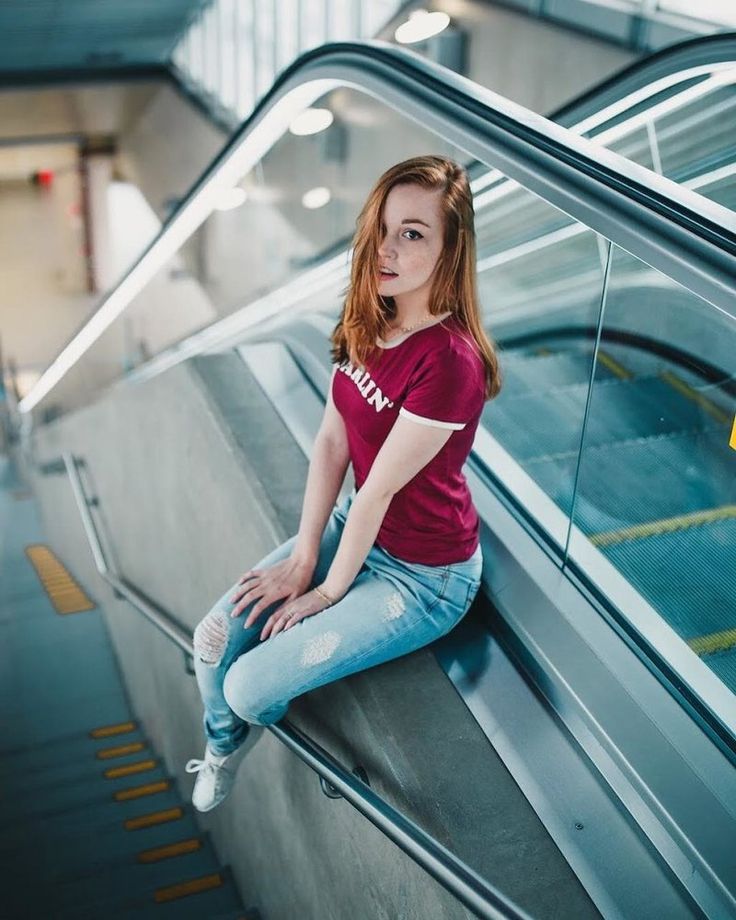 young woman with long red hair is sitting on the side of an escalator, looking directly at the camera. she is wearing a red tshirt and ripped blue jeans. the escalator is made of metal and glass and is located in a modern building. the lighting is natural and coming from the windows above. the perspective is from a high angle, looking down at the woman.