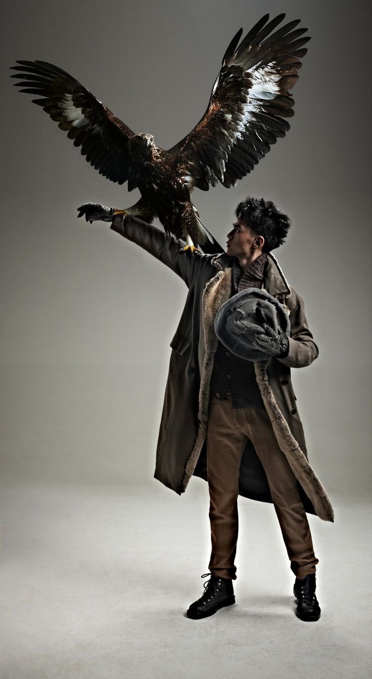 a man in a brown leather coat and fur trimmed collar is posing while holding a golden eagle, the man is looking away from the camera, the studio backdrop is gray, the man is wearing black leather boots, his pose is cool and sharp, and he looks very confident.