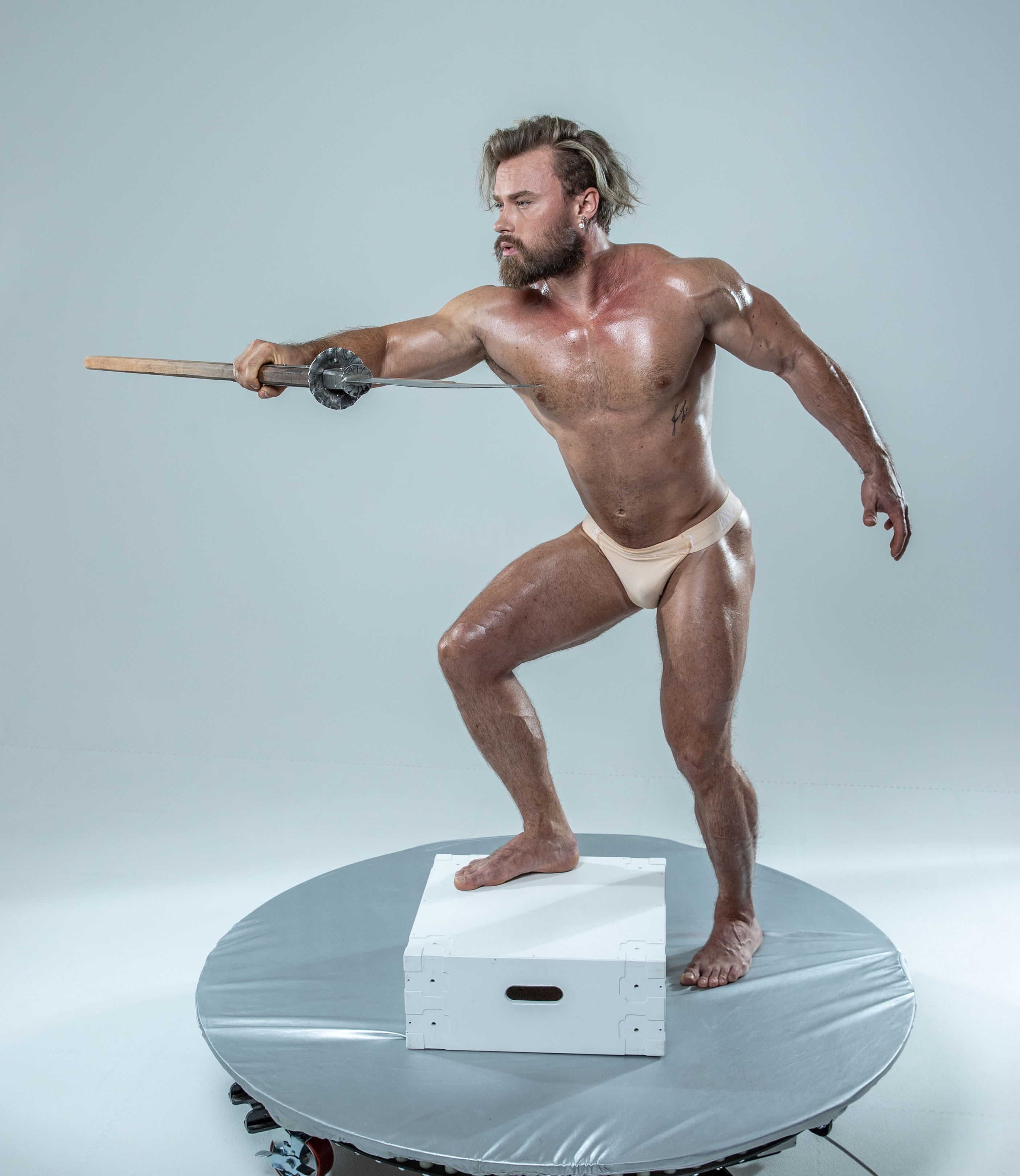 a muscular, bearded man with long blond hair is captured midaction in a studio setting. he stands on a white platform, positioned against a light gray backdrop. his body is tensed in a dynamic pose as he wields a sword, the blade extending towards the left.  his focused expression and powerful physique convey a sense of strength and readiness for battle. the studio lighting highlights his muscular definition and the gleam of the sword.