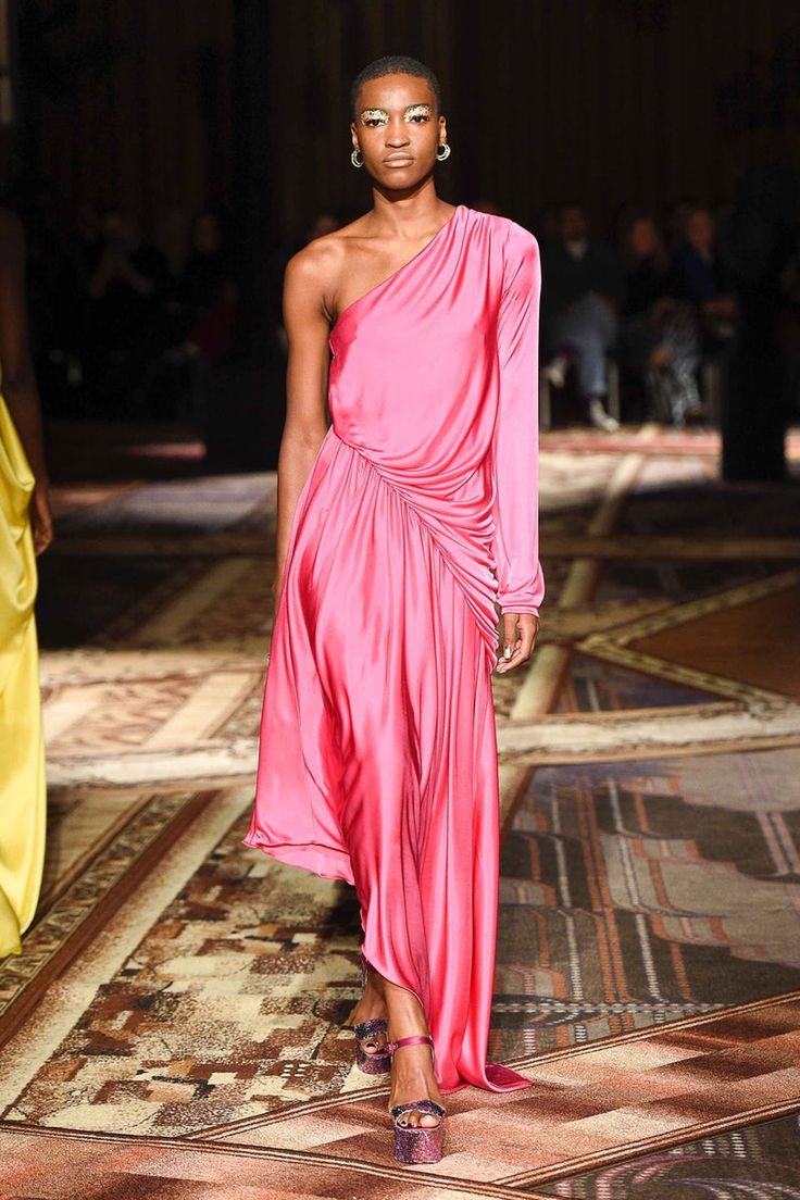 a fashion model walks down a runway wearing a vibrant pink, oneshoulder, longsleeve silk dress. the dress is draped and flows gracefully to the floor. she is wearing platform high heels and has short hair with minimal makeup and earrings. the background shows a patterned carpet typical of fashion show venues, with blurred spectators in the distance. the overall image is one of elegance and sophistication in the fashion industry.