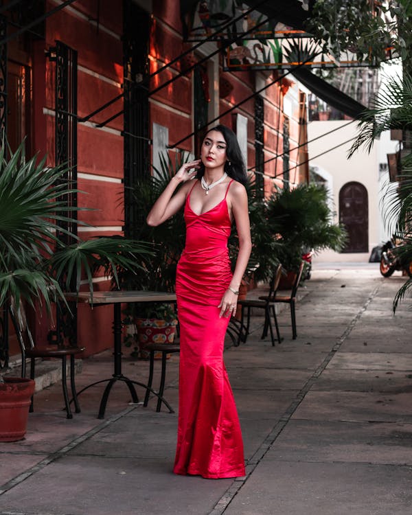 a fulllength shot of a young woman wearing a vibrant red satin mermaidstyle gown. she is standing confidently in a charming, narrow alleyway with colonialstyle buildings on either side.  tropical plants and greenery frame her, adding to the overall ambiance. the woman has dark hair and is wearing a delicate necklace.  the setting appears to be in a latin american city, possibly mexico, given the architecture. the overall mood is elegant and romantic.