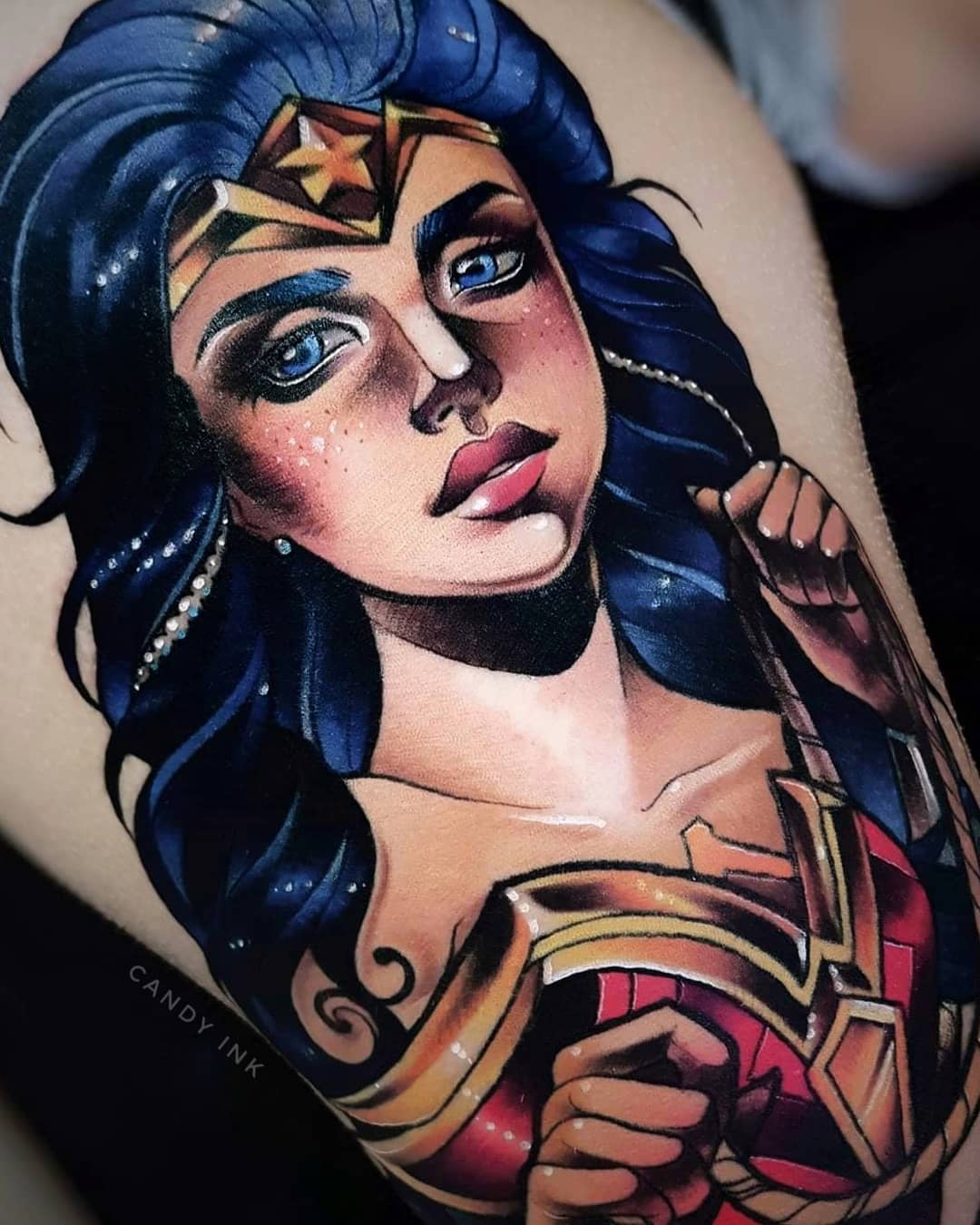 a colorful tattoo of wonder woman on someone's upper thigh. the tattoo is a portrait of the superhero with blue eyes, red lips, and her signature gold tiara. she is wearing her red and gold bodice and has her arm raised. the tattoo is very detailed and wellexecuted.