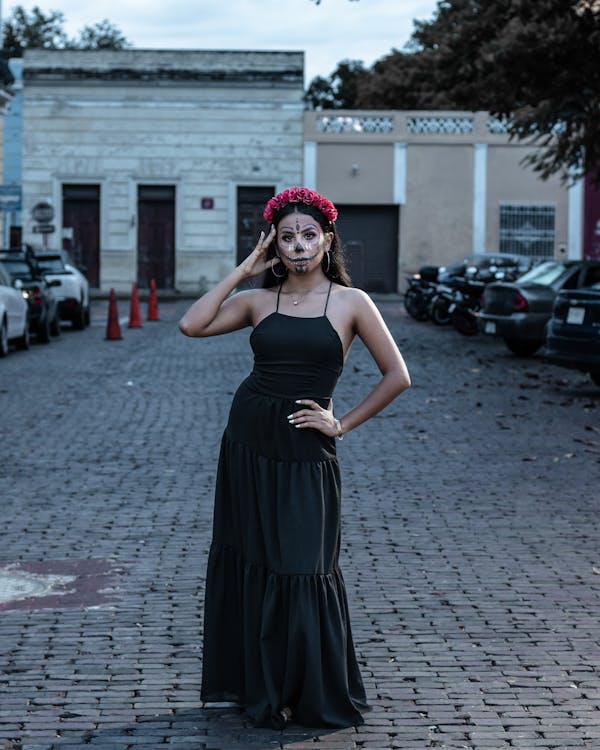 a woman wearing a sugar skull makeup look and a floral crown stands on a cobblestone street in an old town. she is wearing a black maxi dress and is posing for a photo. cars are parked on either side of the street. the overall mood is autumnal.