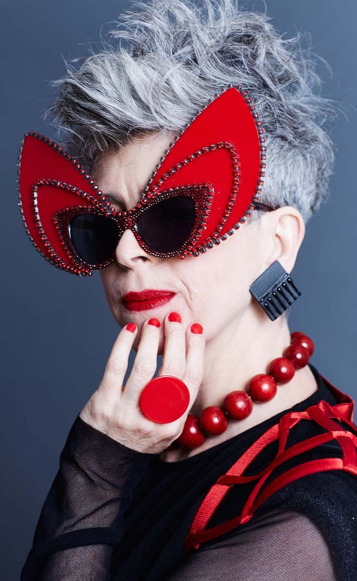 loseup portrait of a stylish older woman with short, spiky grey hair. she is wearing striking red sunglasses with a unique design, a red necklace, a large red ring, and black earrings. her makeup includes red lipstick. she's dressed in a black top with red accents. the overall look is bold, fashionable, and confident. the background is a simple, dark grey.