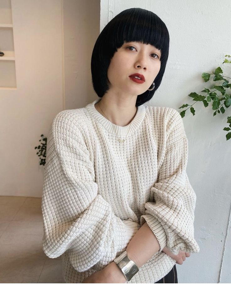 a young woman with short black bob haircut is smiling at the camera. she is wearing a white waffle knit sweater and a silver bracelet.