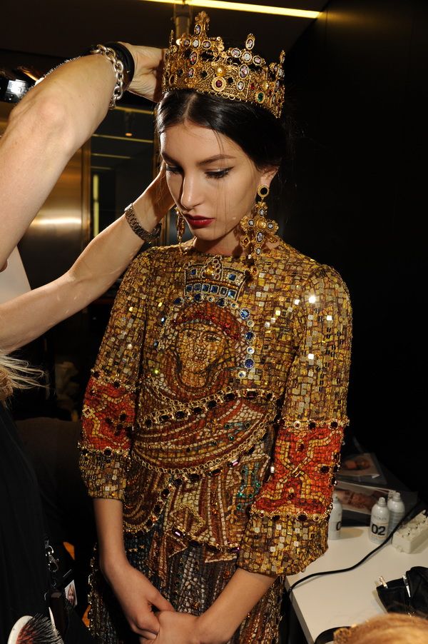 a young woman, possibly a model, is seated backstage at a fashion show.  she's wearing a stunning, intricately beaded gold dress adorned with mosaiclike patterns and a large, ornate gold crown.  another person is placing the crown on her head. the dress is incredibly detailed and luxurious. the overall setting suggests a highfashion event.