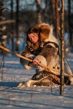 a woman wearing a large fur coat kneels in the snow of a wintery woods, holding a bow and arrow.