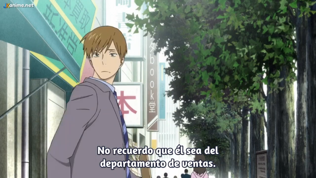 the image shows a young man with brown hair and brown eyes. he is wearing a gray suit and a blue tie. he is walking on the sidewalk on the left side of the image. the street is lined with trees and buildings. there are signs on the buildings, some of which are in japanese. the image is brightly lit, as if it is daytime. the overall tone of the image is neutral.