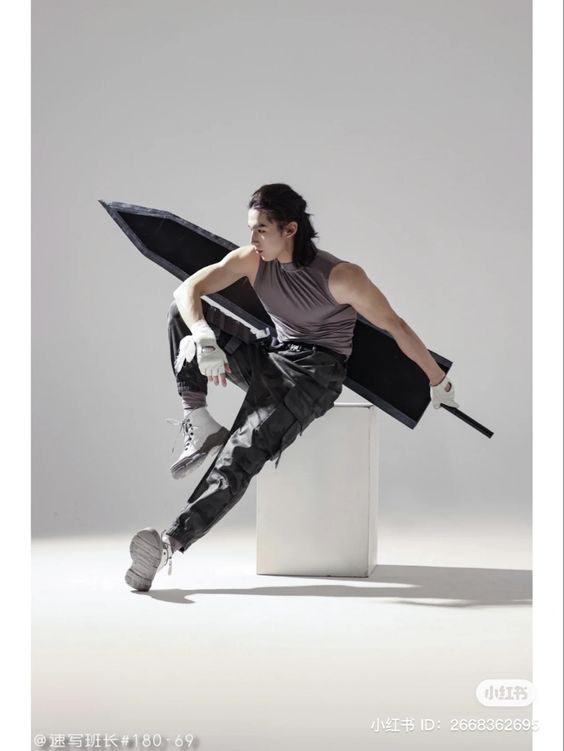 a man is posing with a large black sword in a studio setting. he's wearing a gray sleeveless shirt, camouflage pants, white sneakers, and white fingerless gloves. he's perched on a white cube, holding the sword horizontally with one hand resting on the blade and the other holding the hilt. the background is a smooth, light gray. the lighting is soft and artificial, evenly illuminating the scene.