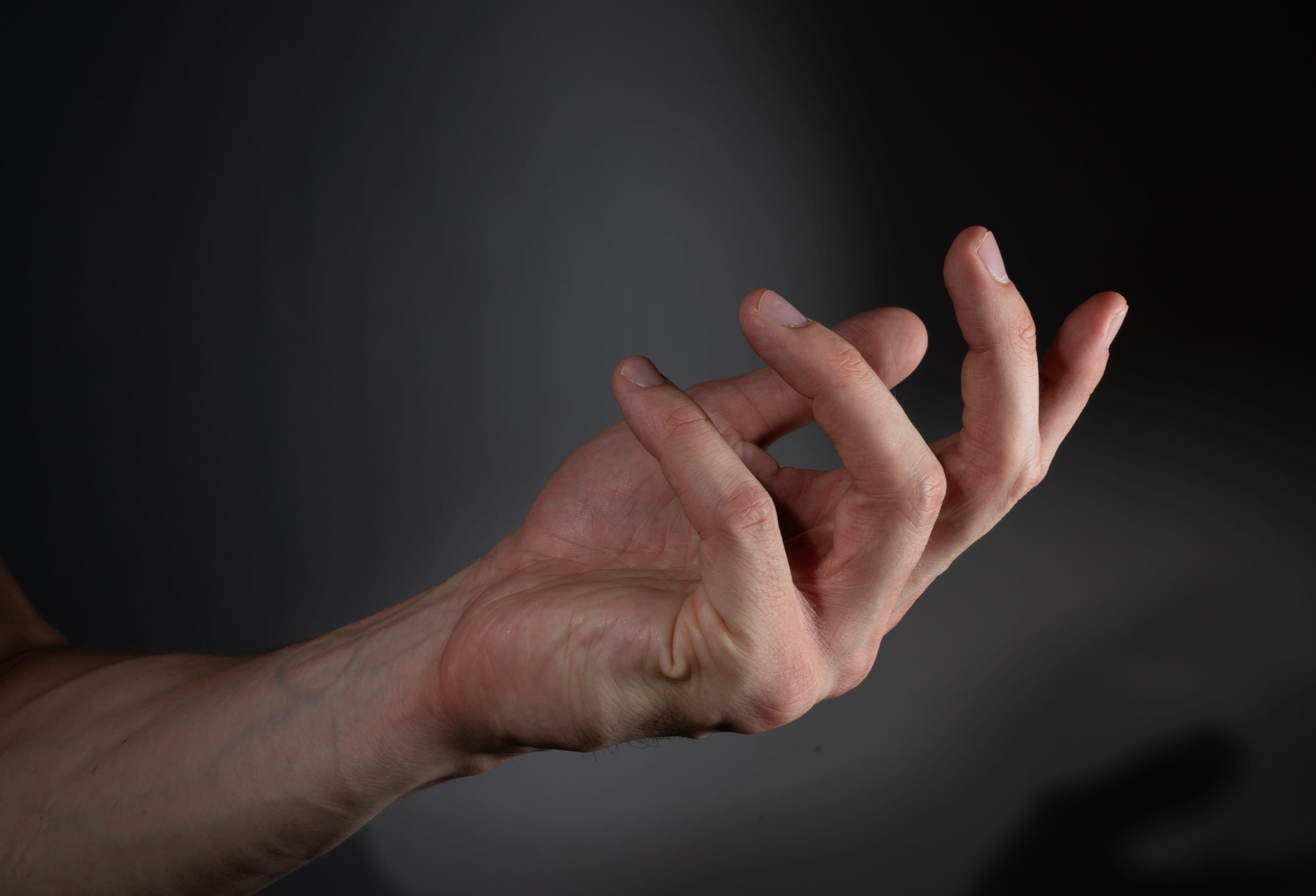 a man's left hand is reaching out against a dark gray background. the image highlights the details of the hand and its tendons, creating a sense of tension or anticipation. the lighting is dramatic, casting shadows that emphasize the contours of the hand.