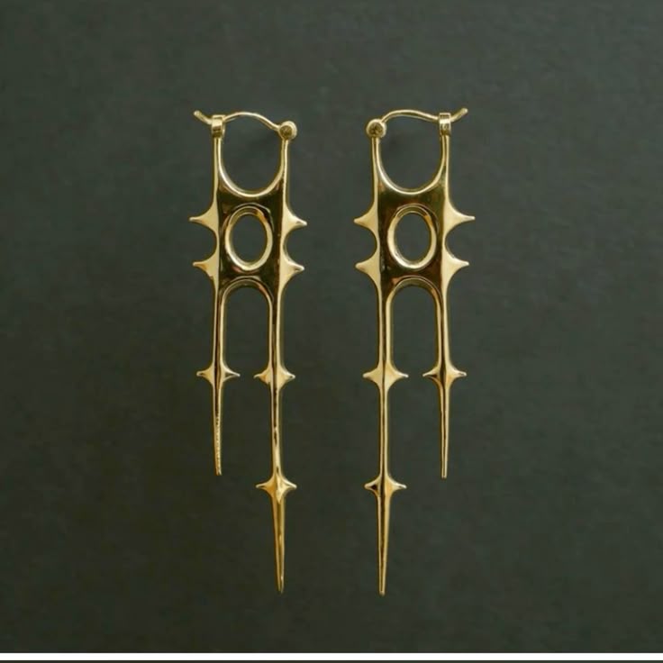 the image shows a pair of gold earrings on a gray background. the earrings are hoop earrings with a unique, spiky design. they dangle and feature a circular element in the middle.