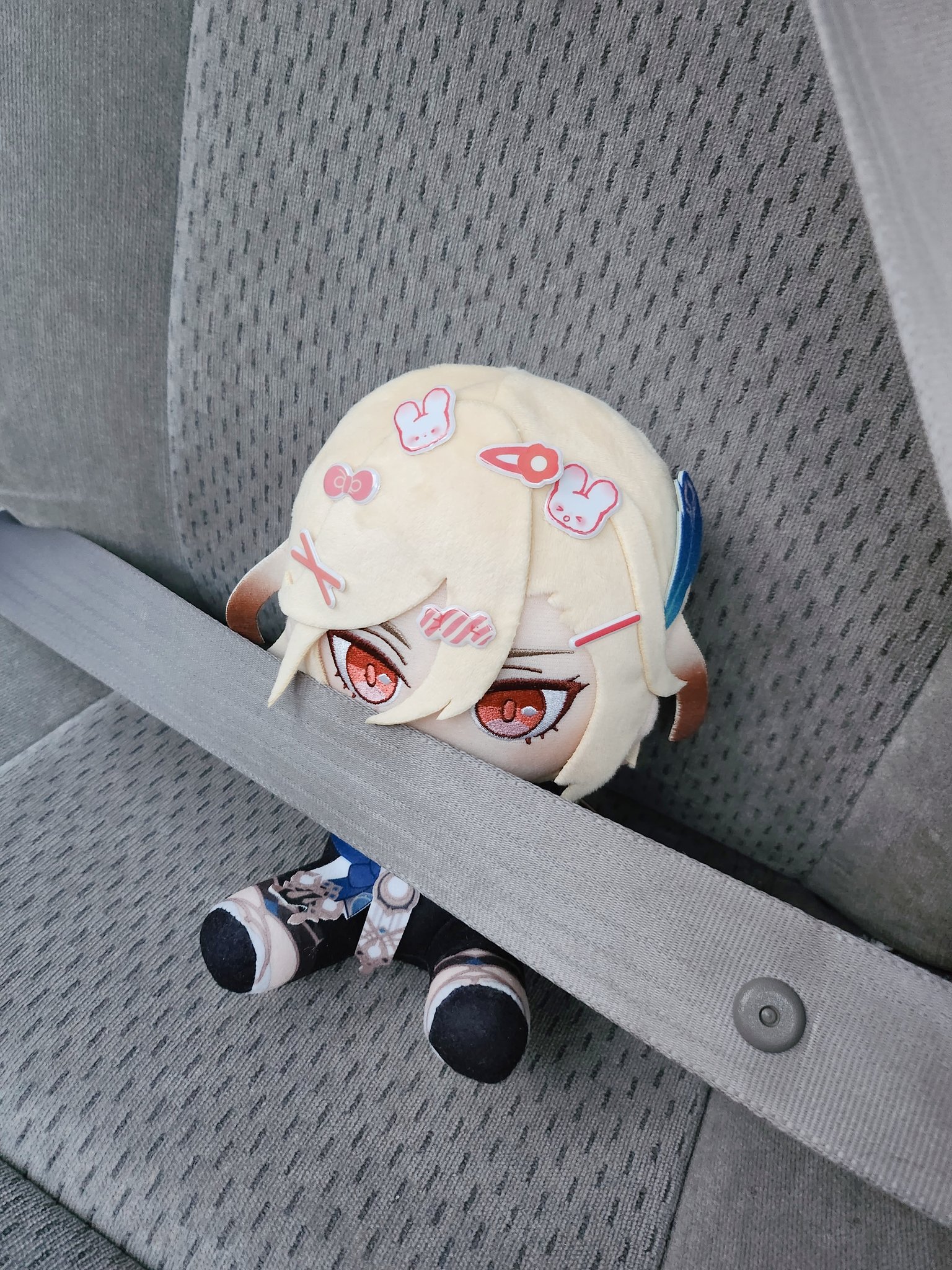 a plush toy of an anime character with blonde hair, pink eyes, and accessories is sitting on a grey car seat, with the seatbelt going over it. the seat is grey and made of a soft fabric with small black lines running across it, resembling a grid. the plush toy is wearing black and white clothing, with pink and white accessories.  the plush toy's eyes are closed and they are looking down. the car seat is made of a grey fabric with small black lines running across it. there is a grey car seat belt running across the plush toy's body. the plush toy is positioned to the left of the image, with the car seat belt going over it.  the car seat is covered in fabric with small black lines running across it, resembling a grid.