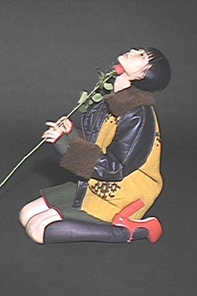 a woman in a black leather jacket, yellow sweater, and green skirt is sitting down on a grey background with a red rose in her hand, she is looking up.