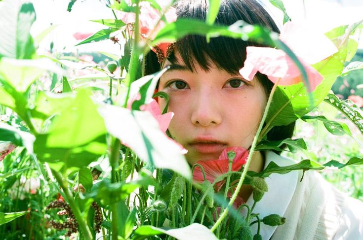 the image is a closeup portrait of a young asian woman's face, partially obscured by lush green plants and pink flowers.  the woman has dark hair and soft features. the flowers are in soft focus, creating a dreamy and ethereal atmosphere. the overall mood is calm and peaceful, suggesting a serene outdoor setting on a bright day. the style of the photo appears to be film photography with a slightly soft focus and natural light. the plants are a significant part of the composition, creating a frame around the woman's face.