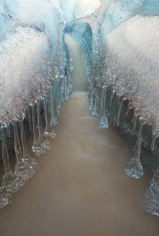the image shows a narrow stream of murky water flowing through an ice cave or glacial tunnel. icicles hang from the icy walls, suggesting melting. the scene depicts the interior of a glacier with light shining through an opening.