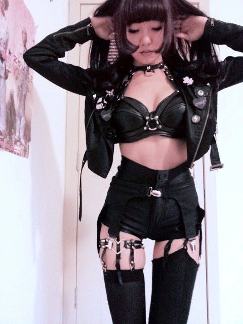 a young woman poses in a black leather jacket, shorts, and garters. her look is very edgy and punkinspired. the photo is taken indoors with a light background.