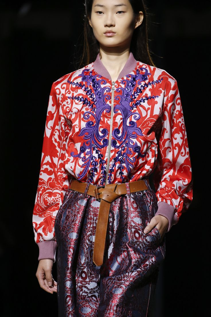 loseup shot of a fashion model showcasing a vibrant autumn/winter outfit. the model, with long dark hair, is wearing a red bomber jacket adorned with intricate purple floral embroidery.  the jacket is paired with a dark, richly patterned skirt or highwaisted pants with a similar design. a brown leather belt cinches the waist. the background is dark and unfocused, highlighting the clothing. the overall aesthetic is elegant and stylish, typical of a runway presentation.