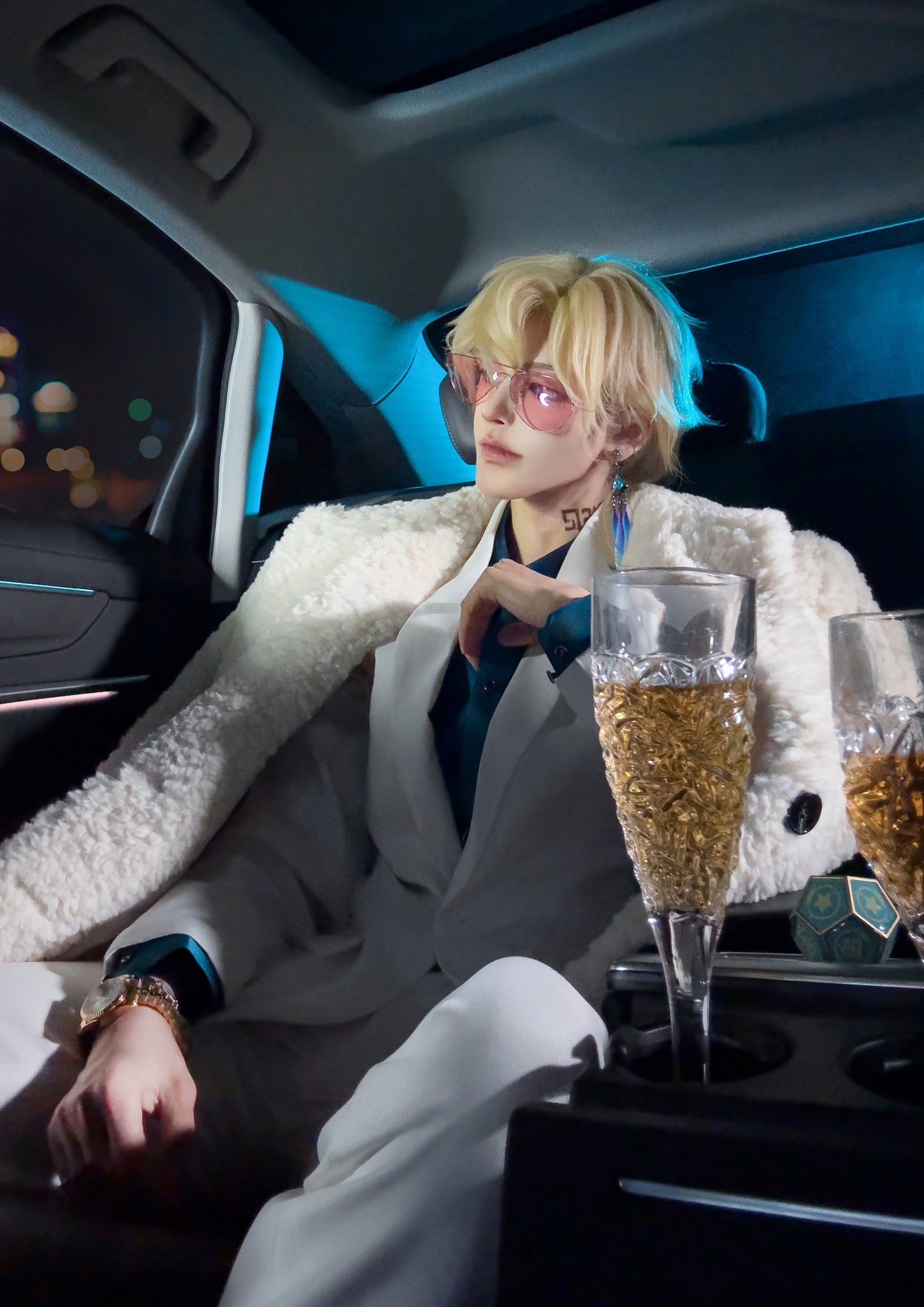 a blonde haired man in a white suit and sunglasses sits in the back seat of a car. he is holding a champagne glass in his right hand and looking out the window. he is wearing a gold watch on his wrist.