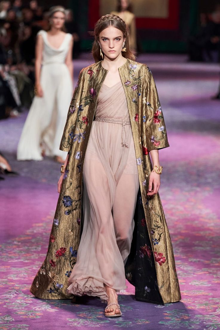 a model walks the runway at a dior haute couture fashion show. she is wearing a long, flowing pink dress underneath a gold, embroidered coat with floral patterns. the coat has threequarter sleeves and falls to the floor. the model's hair is styled in a braid, and she is wearing minimal makeup. she accessorizes with sandals and gold jewelry. the runway is purple, and other models can be seen in the background.