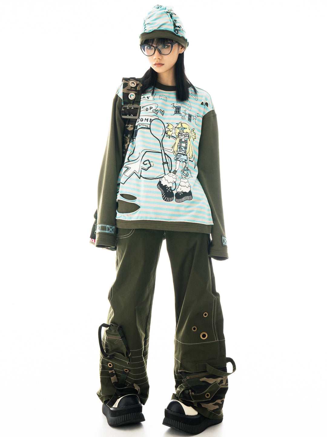 a full shot of a female model posing in studio against a white background. she's wearing a light blue and white striped longsleeve shirt with an animeinspired graphic, olive green cargo pants with added camouflage details, black combat boots, and a green beanie. the style is a mix of streetwear, punk, and alternative fashion with a distinct japanese influence.  the shirt shows distressing or rips. the overall aesthetic is edgy and stylish.