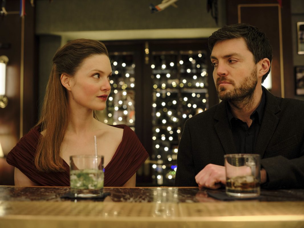 the image shows a man and a woman sitting at a bar, seemingly on a date.  the woman is wearing a darkred dress, and the man is wearing a dark suit.  they are looking at each other, and each has a drink in front of them: the woman appears to have a gin and tonic, and the man has something darker, possibly whiskey. the bar setting is dimly lit and atmospheric, with a blurred background of warm lights creating a bokeh effect. the overall mood is romantic and intimate, suggestive of a scene from a film or television show. the style is elegant and stylish.