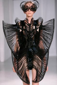 a female model is wearing a striking black and white ensemble. the dress is a complex and futuristic design, composed of geometric, structured, and feathery elements. the dress resembles wings, creating a dramatic silhouette. the model is wearing a pair of large, black,  feathery glasses that add to the avantgarde feel of the outfit. this striking look is a testament to high fashion's innovative and artistic nature.
