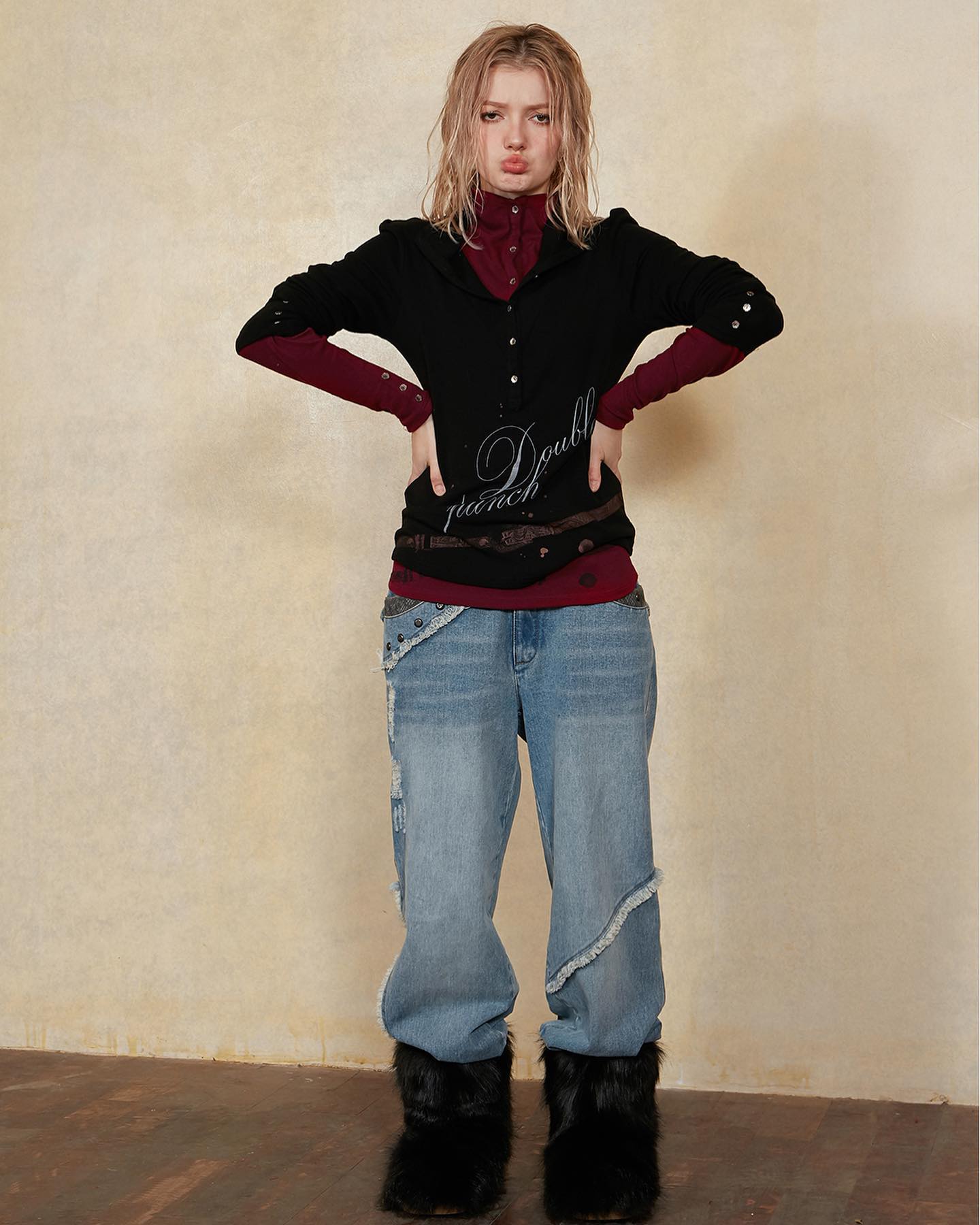 a full shot of a young woman posing in a studio. she is wearing a black longsleeved sweater with a burgundy undershirt, light blue distressed jeans, and black furry boots. her blonde hair is loose. the background is a light beige wall. the overall style is casual and trendy, with a hint of grunge or alternative fashion.