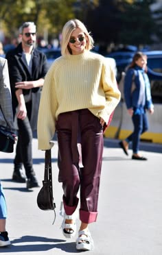 a stylish woman walks down the street wearing a casual yet chic outfit. she is wearing a pale yellow sweater, burgundy leather pants, and white sneakers. she is accessorizing with sunglasses and a black bag. she looks confident and stylish as she walks along the city streets.  the outfit is casual yet chic and would be perfect for a day of running errands or a night out with friends.