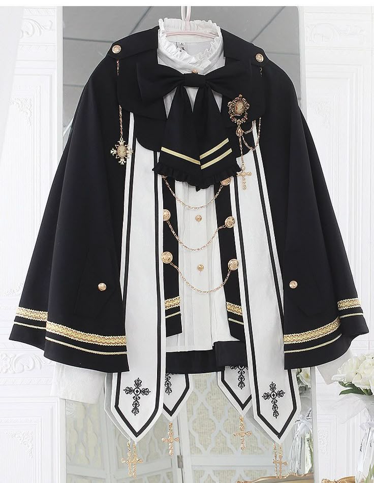 a black and white lolita fashion cape hangs on a hanger, displaying intricate details like gold accents, a bow, crosses, and a decorative brooch. hanging crosses adorn the bottom, adding to its ornate and formal design.