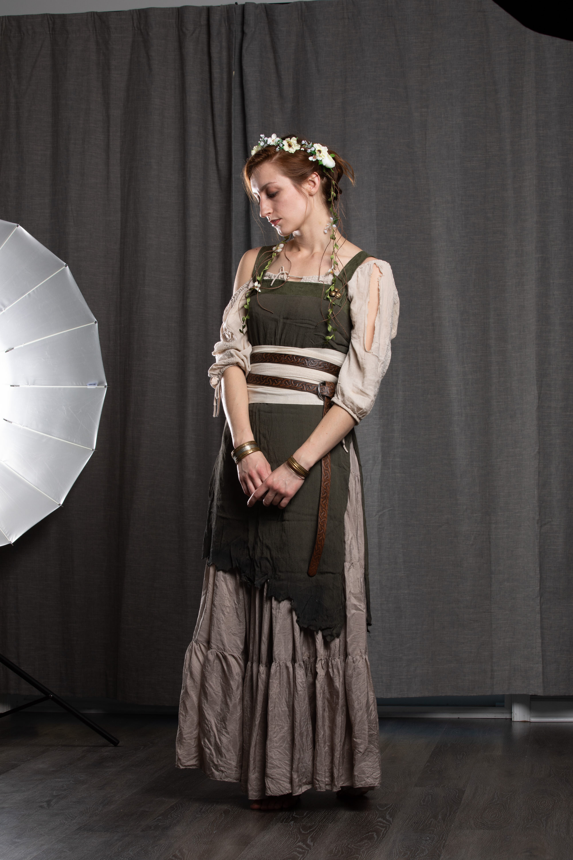 a young woman with reddishbrown hair, wearing a floral crown and a flowing, layered medievalinspired dress, stands against a dark grey backdrop in a photography studio. she has a contemplative expression and her hands are clasped in front of her. the dress is primarily olive green and beige, with a leather belt and accents. the studio is minimally lit with a single softbox, creating soft and diffused lighting.