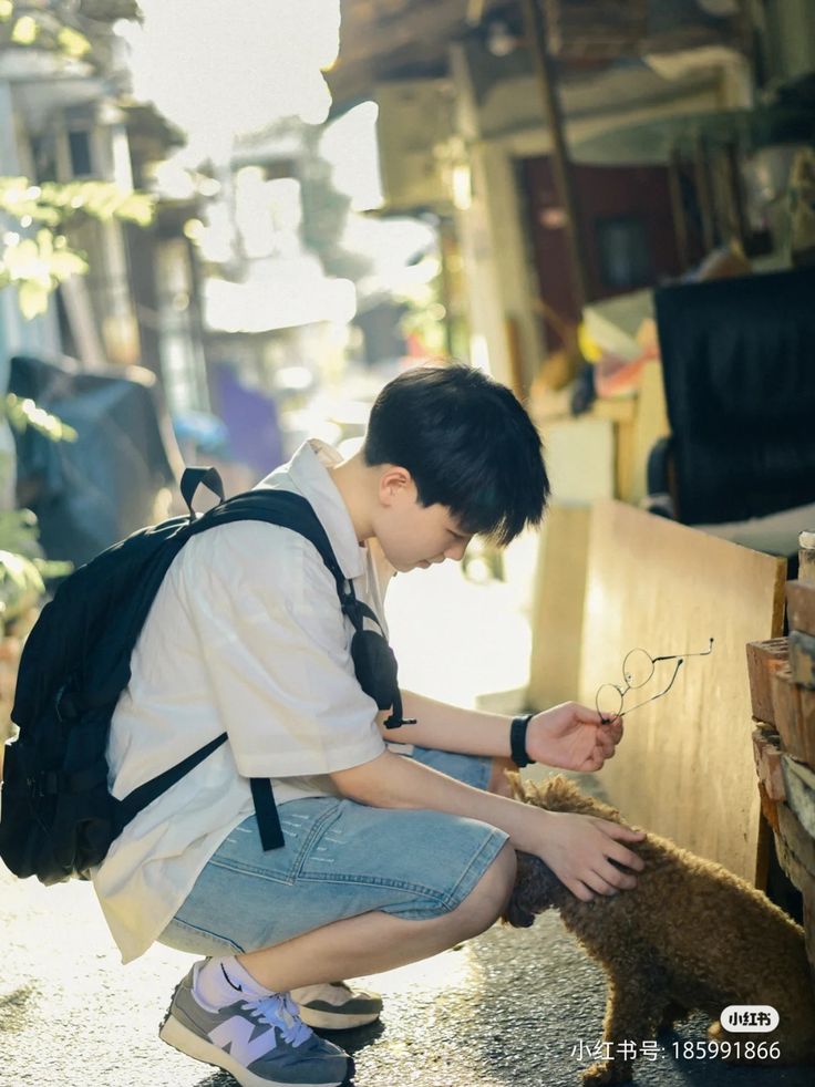 a young man wearing a white shirt and denim shorts is crouching down in a narrow alleyway, petting a small brown dog. he has a backpack slung over one shoulder and is holding a pair of glasses in his other hand. the scene is bathed in warm, natural light, creating a gentle and candid atmosphere. the background suggests an urban environment with glimpses of buildings and various objects.
