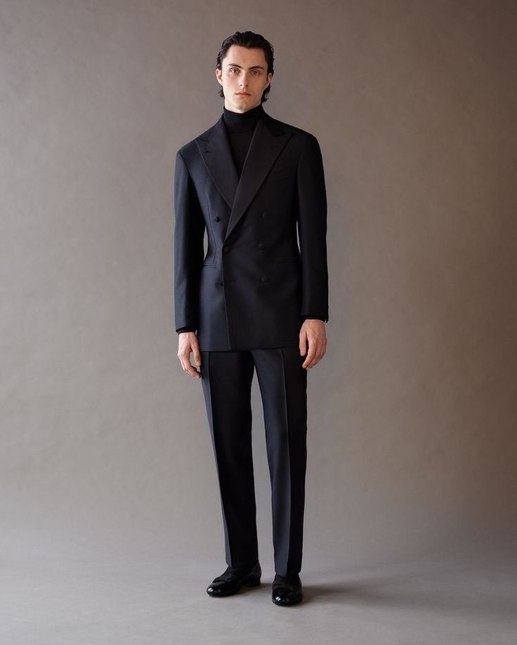 a fulllength studio shot of a man wearing a black doublebreasted suit with a black turtleneck. he is standing with his arms at his sides, looking directly at the camera. he also wears black dress shoes. the background is a neutral greybrown color. the suit is a slim fit and appears to be tailored.