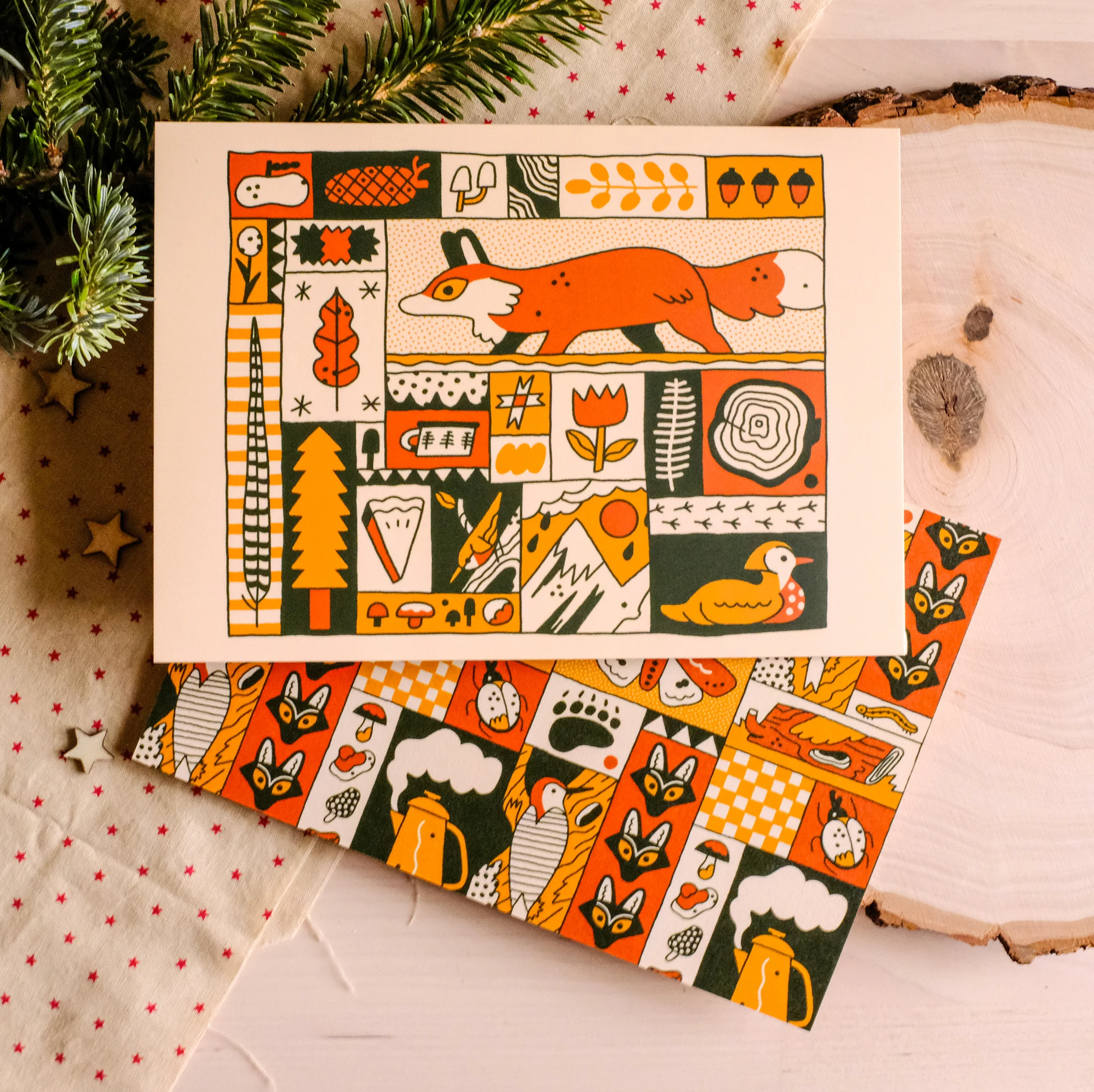 a flatlay photograph shows a christmas card featuring a whimsical illustration of a fox in a forest setting.  the illustration is composed of various patchwork squares with different naturethemed elements. the card rests on a piece of fabric with a subtle star pattern, near a wood slice, and on top of matching wrapping paper. a fir branch is slightly visible in the top left corner, adding to the holiday theme.  the overall style is cute, adorable, and whimsical.