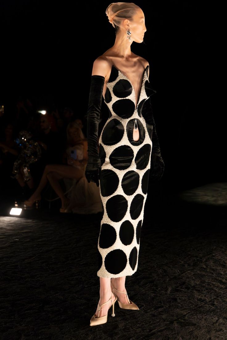 a model on a runway, wearing a black and white polka dot dress with black gloves and beige heels. the dress has a deep vneck and a small pink handbag attached to one of the polka dots. the model is wearing a nudecolored head covering and silver earrings. the background is dark, with blurred figures in the distance. the runway is black.