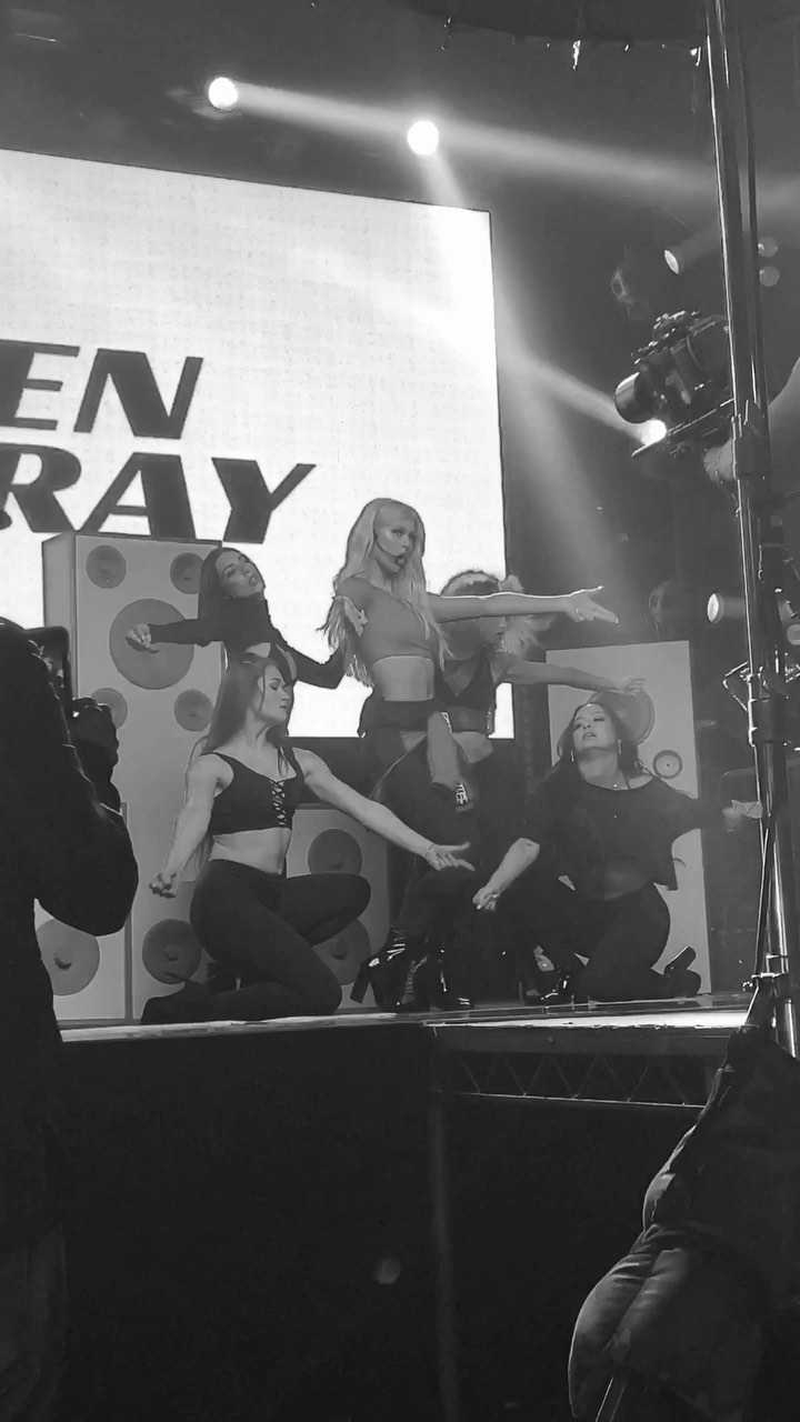 a black and white photograph shows a singer performing on stage with backup dancers in front of a large screen with text that appears to say "en ray". the singer, with long blonde hair, is center stage with several female dancers around her, all posed in what appears to be a choreographed routine. the lighting is focused on the performers, and the image suggests an active, energetic concert scene. there is recording equipment on the right.