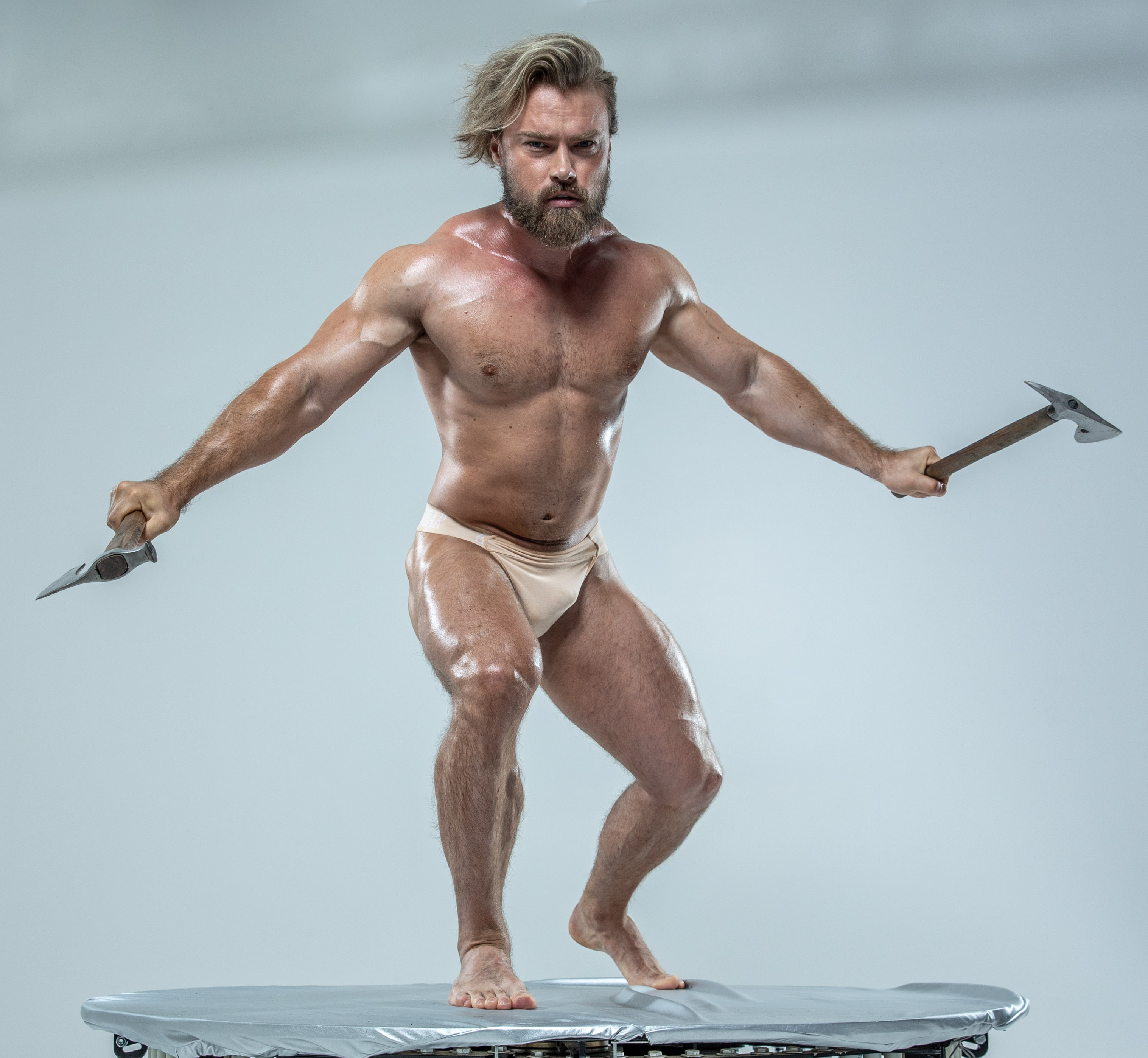 a muscular man with an axe in each hand is standing on a round platform in a studio setting. he is wearing only underwear and has a beard and long blond hair. the background is white and the lighting is soft. he is holding the axes up in a fighting stance.