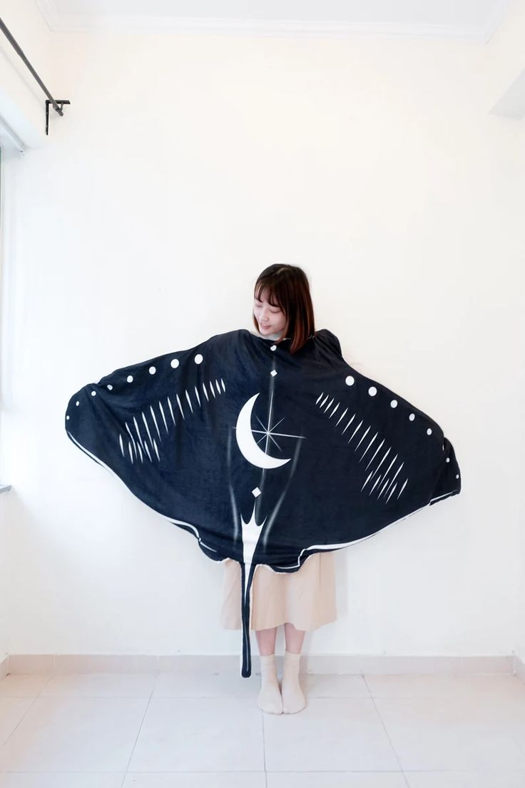 a woman wearing a black and white moon blanket, posed with her hands at her side and looking down. the blanket is in the shape of a large manta ray.  the woman is standing in an empty room with white walls and tiled floors.