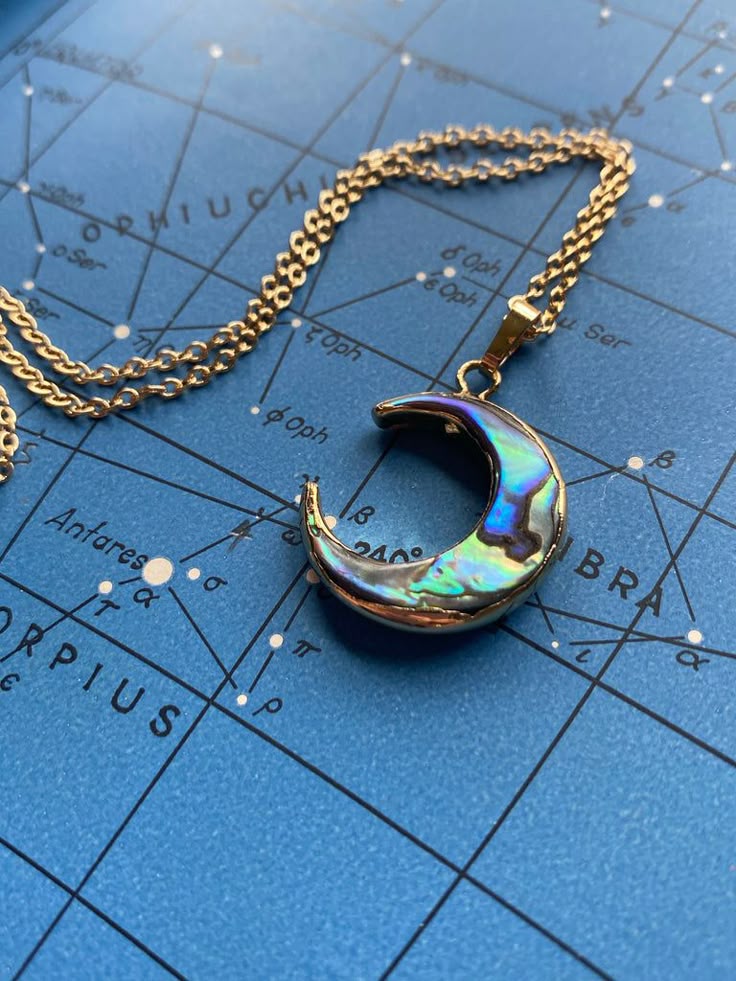 a closeup shot of a gold moon pendant necklace lying on a blue background with constellation map details visible. the moon pendant is iridescent and the chain is a delicate gold link.