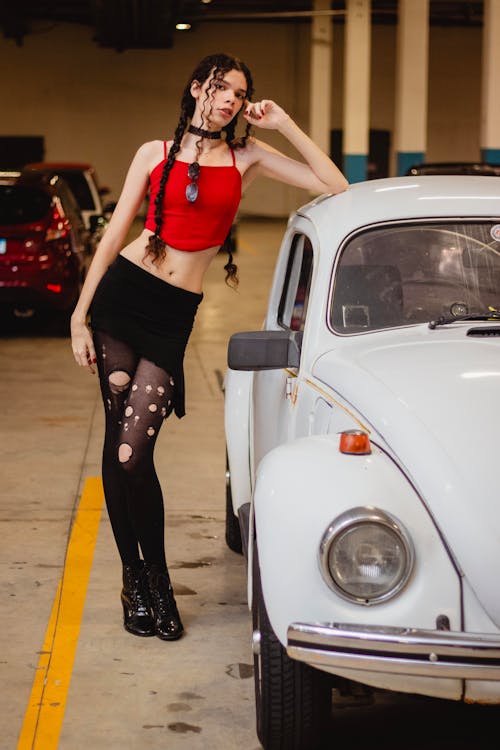 a young woman with curly hair in braids is posing next to a white vintage volkswagen beetle in a parking garage. she is wearing a red crop top, black skirt, and ripped black tights, along with black ankle boots.  she has a choker necklace and sunglasses around her neck.  the overall style is casual yet fashionable. the image is a full body portrait, with the woman and car as the main focus. the background is slightly blurred, drawing attention to the subject.