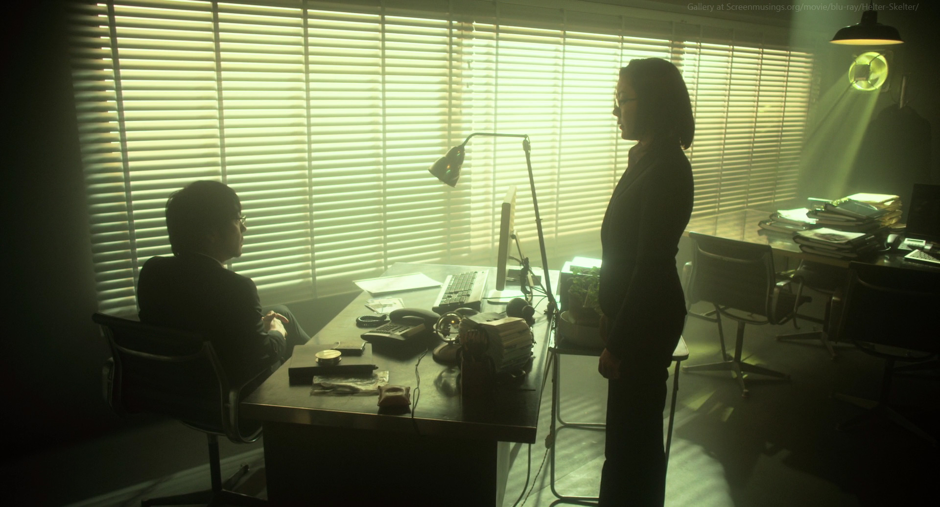 a man and a woman are having a meeting in an office. the man is silhouetted in the chair, looking towards the woman. the woman is standing, looking towards the man. the office has blinds on the windows, a desk with a computer, a lamp, and chairs. there is a large amount of papers stacked in the background.