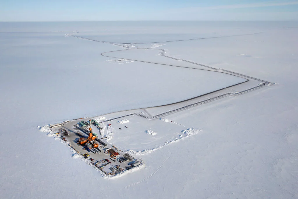 an aerial view of a drilling rig in the arctic, surrounded by snow and ice. the rig is located on a remote, barren landscape. the image captures the harsh conditions of the arctic and the scale of oil and gas exploration in the region.