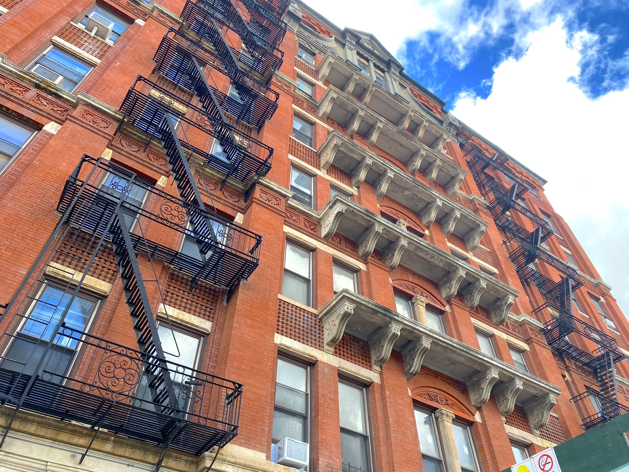 this is a low angle view of an old, red brick apartment building, likely in new york city. the facade is detailed with stone cornices above each row of windows and decorative brickwork. multiple black metal fire escapes zigzag up the front of the building. the image is taken in natural light and the sky is bright blue with puffy white clouds.