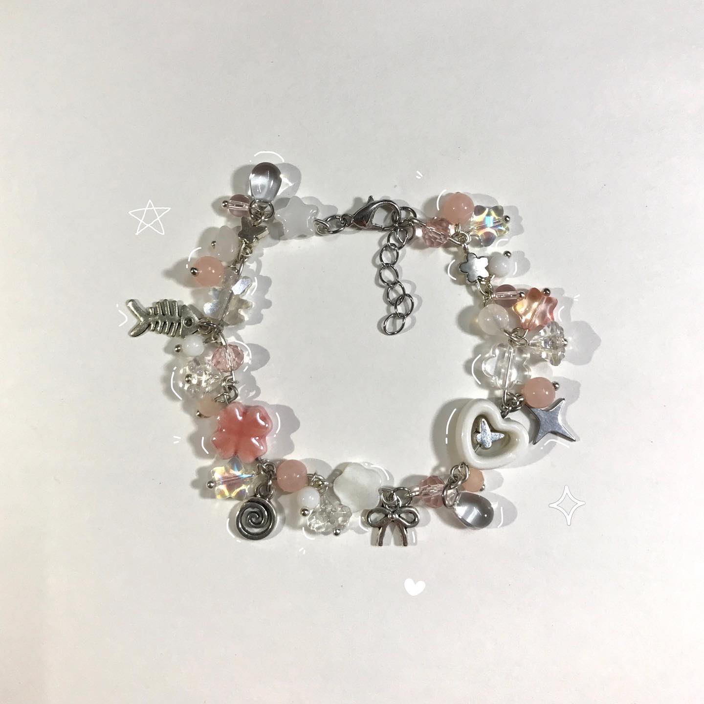 the image shows a charm bracelet with various beads and charms in pink, white, and silver colors. the charms include a fishbone, a flower, a spiral, a bow, a heart, and a star.