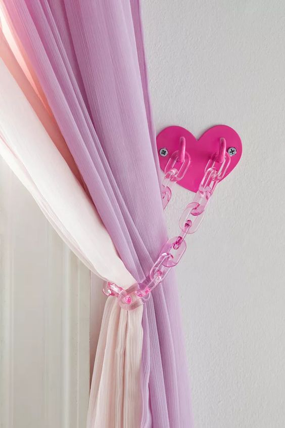a closeup image showing pink sheer curtains tied back with a plastic chain and heart shaped hooks on a white wall. the curtains are a gradient of pink to white and add a touch of girly decor to the room.