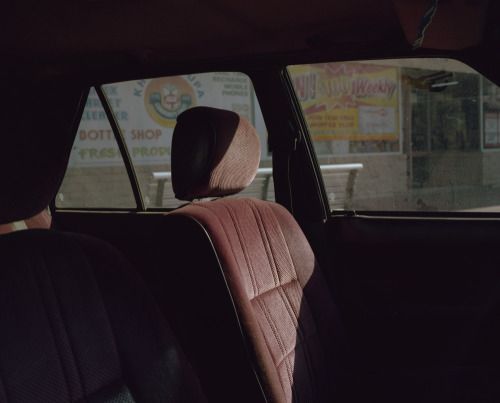the image shows the interior of a vintage car, likely from the 1980s, focusing on the back seats. the seats are a deep maroon color with vertical ribbed upholstery. the headrests are also maroon and upright. the car is parked, and the side windows show a reflection of a street scene with a shop visible; a brightly colored sign is partially visible in the window.  the overall lighting suggests it's daytime, with sunlight illuminating the interior of the car. the car appears empty, giving a sense of quiet stillness. the focus is primarily on the textures and colors of the car's interior.